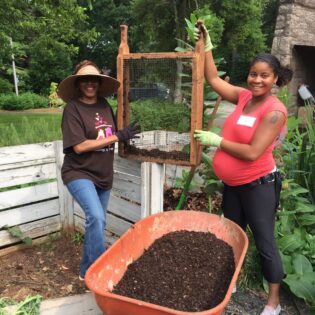 Two women sieve fresh compost into a wheelbarrow. They work in landscaped clearing framed with lush woods.