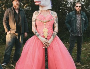 A frontwoman in a pink princess gown and unicorn mask holds a painted banjo. She is flanked on each side by her 2 bearded male bandmates in flannel shirts and jeans. This is Heather and the Possumden.