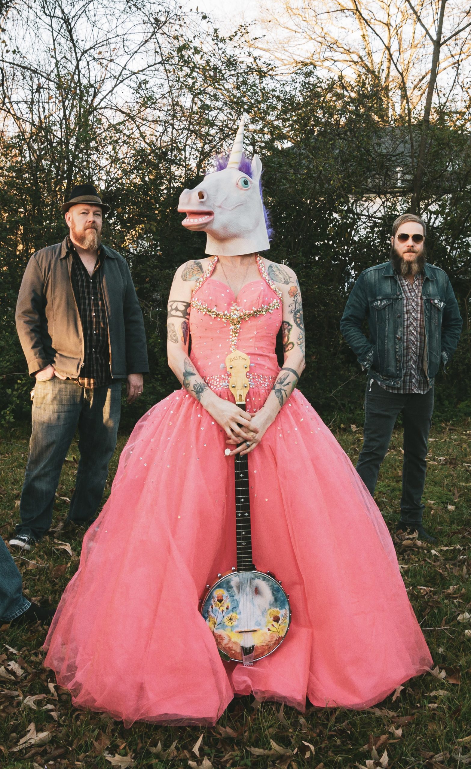 A frontwoman in a pink princess gown and unicorn mask holds a painted banjo. She is flanked on each side by her 2 bearded male bandmates in flannel shirts and jeans. This is Heather and the Possumden.