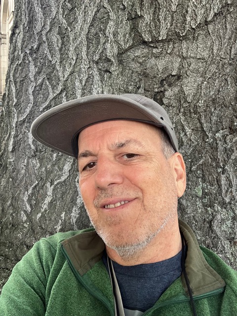 Neil Norton, ISA certified master arborist, is pictured in a green pullover and field cap against the grey and deeply furrowed bark of a wide tree.